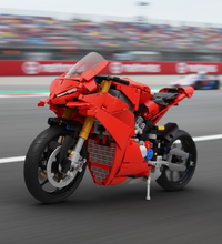 A red brick-built toy motorcycle, resembling a sportbike, constructed from interlocking plastic blocks. It features a detailed design with black wheels, gold forks, and silver accents, shown on a neutral gray surface with a blurred white background containing a red sphere.