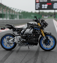 Two views of a toy building-block motorcycle on a plain light gray background. The motorcycle is primarily black and gray with blue wheel accents and gold front fork and chainring details.