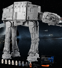 A large gray toy AT-AT walker built from interlocking blocks, shown on a white background with small accessory figures, a black box, and an orange piece set at the bottom.