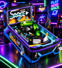 A toy building-block pinball machine themed around space, shown open to reveal a detailed playfield with planets, stars, and colorful retro game elements on a plain white background.