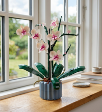 A building-block model of a potted orchid plant, featuring white flowers with pink centers, dark green leaves and stems, and a gray pot, shown on a plain white background with an orange accessory piece at the bottom left.