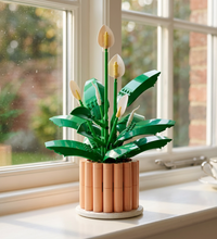 A building-block style plant arrangement with glossy green leaves, white spatelike flowers, yellow centers, and a peach-colored cylindrical pot on a plain white background.