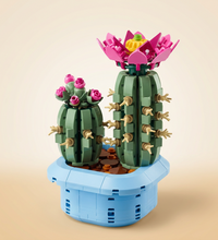 A toy building-block style cactus arrangement in a light blue planter, with two green cactus shapes, pink flowers, tan spines, and a yellow flower center on a plain white background.