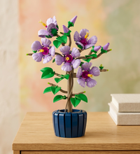 A LEGO-style assembled flowering tree in a dark blue ribbed pot, shown on a plain white background. The flowers are primarily light purple/lavender with magenta centers, green leaves, brown branches, and small yellow accents.
