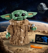 A built Star Wars The Child (Grogu) set with a brown robe, green skin, and accessories including a black display plaque, a small figure, and an orange accessory, shown on a white background.
