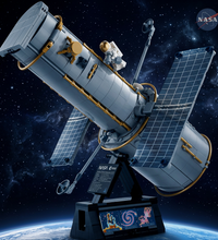 A detailed model of a space station or satellite on a black display stand, featuring gray cylindrical sections, blue solar panels, gold accents, and a small astronaut figure, shown against a plain white background.