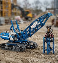 A blue and white brick-built construction crane toy with black tracks, shown alongside a matching blue support frame on a plain white background.