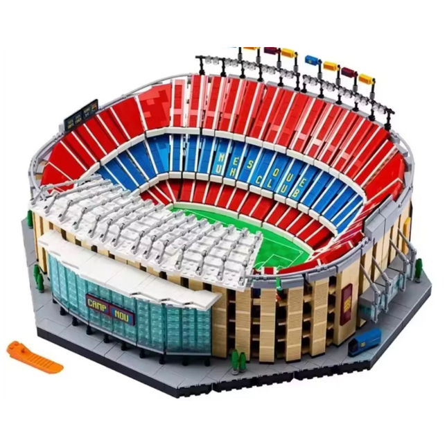 A detailed toy model of a stadium built from interlocking blocks, featuring red and blue tiered seating, a green field, beige stands, and a gray base. An orange accessory piece is placed in the lower-left corner on a plain white background.