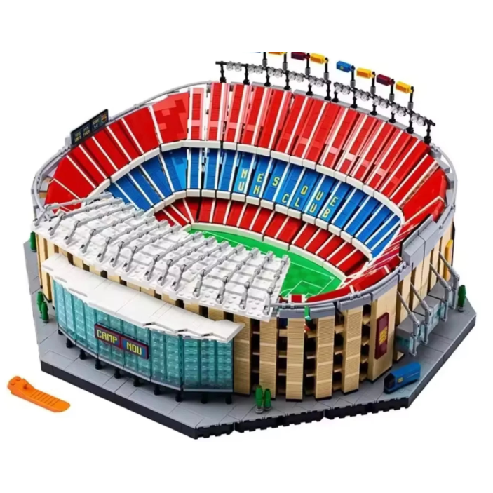 A detailed toy model of a stadium built from interlocking blocks, featuring red and blue tiered seating, a green field, beige stands, and a gray base. An orange accessory piece is placed in the lower-left corner on a plain white background.